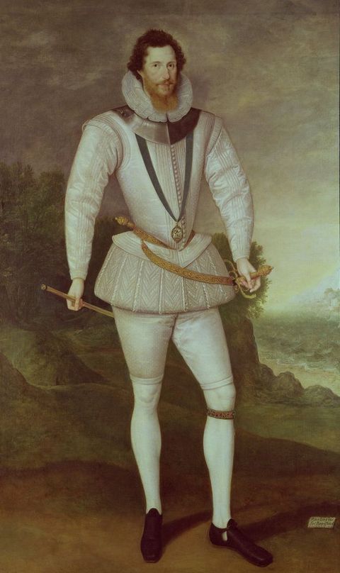 Painting. A pale-skinned man wearing a white suit stands in front of a landscape and looks at us. His brown curls are brushed back from his forehead, and a long ginger beard rests in the pleated ruff around his neck. A gorget, a piece of armour worn around the neck, is over his white jerkin. A sword hangs from a skirt-like, puffed breeches around his hips. His legs are clad in white stockings.