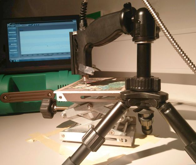 A machine with an arm on a tripod holds a pointed tip just over the surface of a work of art. A computer screen is beyond.